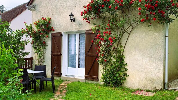 A patio with a table and chairs, a small garden with a brick path, and a building with a window and shuttered door.