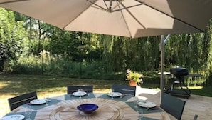 A round wooden table with a central bowl, surrounded by chairs and plates, under a large umbrella. - Charming Retreat in Berry: Heated Pool, Canoeing, Fishing, Near George Sand's House (Ardentes)