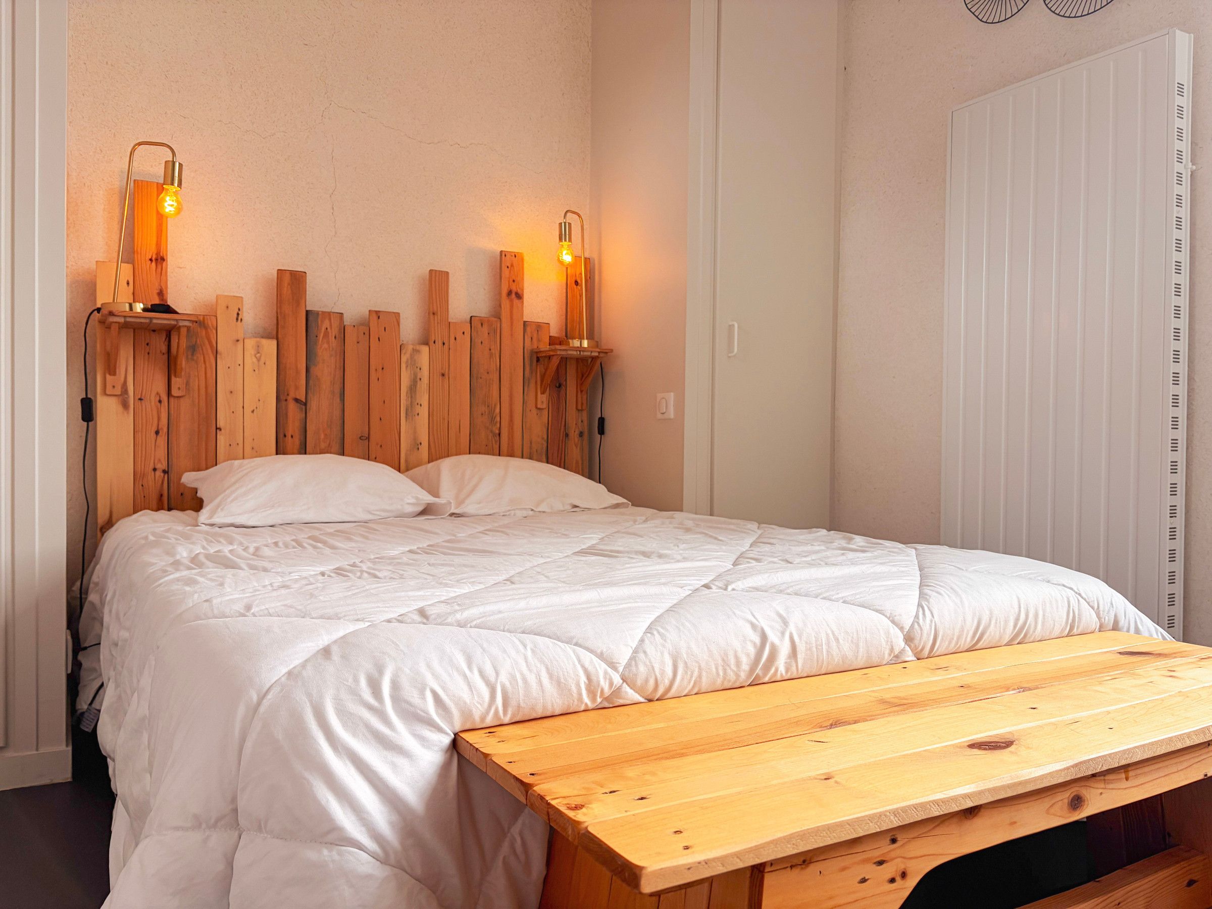 A bed with a wooden headboard, two wall-mounted light fixtures, and a wooden bench.