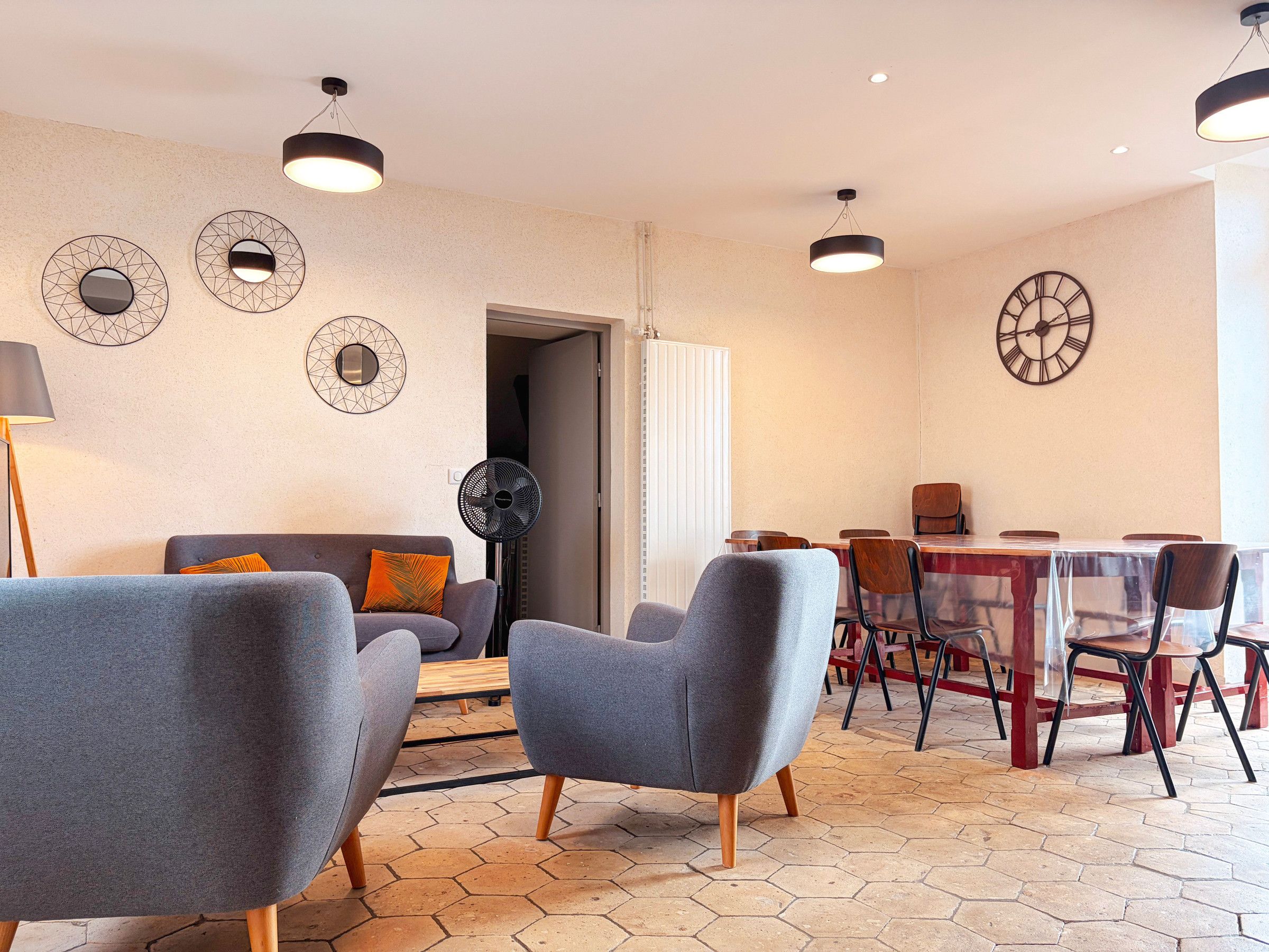 Un salon moderne avec des fauteuils gris, un canapé, une table à manger avec des chaises et des horloges décoratives au mur.