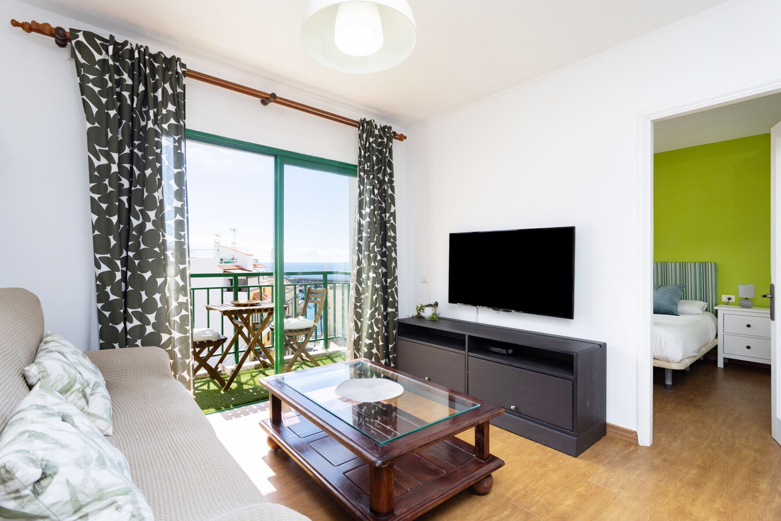 Ein modernes Wohnzimmer mit einem Glastisch, einer Couch, einem Flachbildfernseher und einem Balkon mit Tisch und Stühlen.