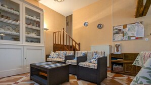 Una sala de estar con muebles de mimbre, piso a cuadros, un armario blanco con vajilla, una escalera y un tablón de anuncios.
