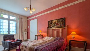 Un dormitorio con cama, una silla, dos mesitas de noche con lámparas, un cuadro en la pared y una ventana con cortinas.