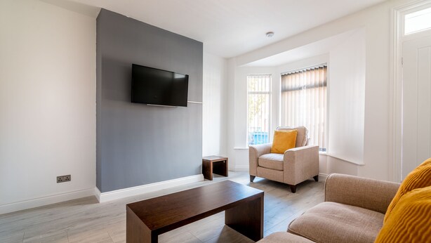 A modern living room with a flat-screen TV mounted on a grey wall, a wooden coffee table, beige sofas, and a small side table. - Platform Avenues House (Hull)
