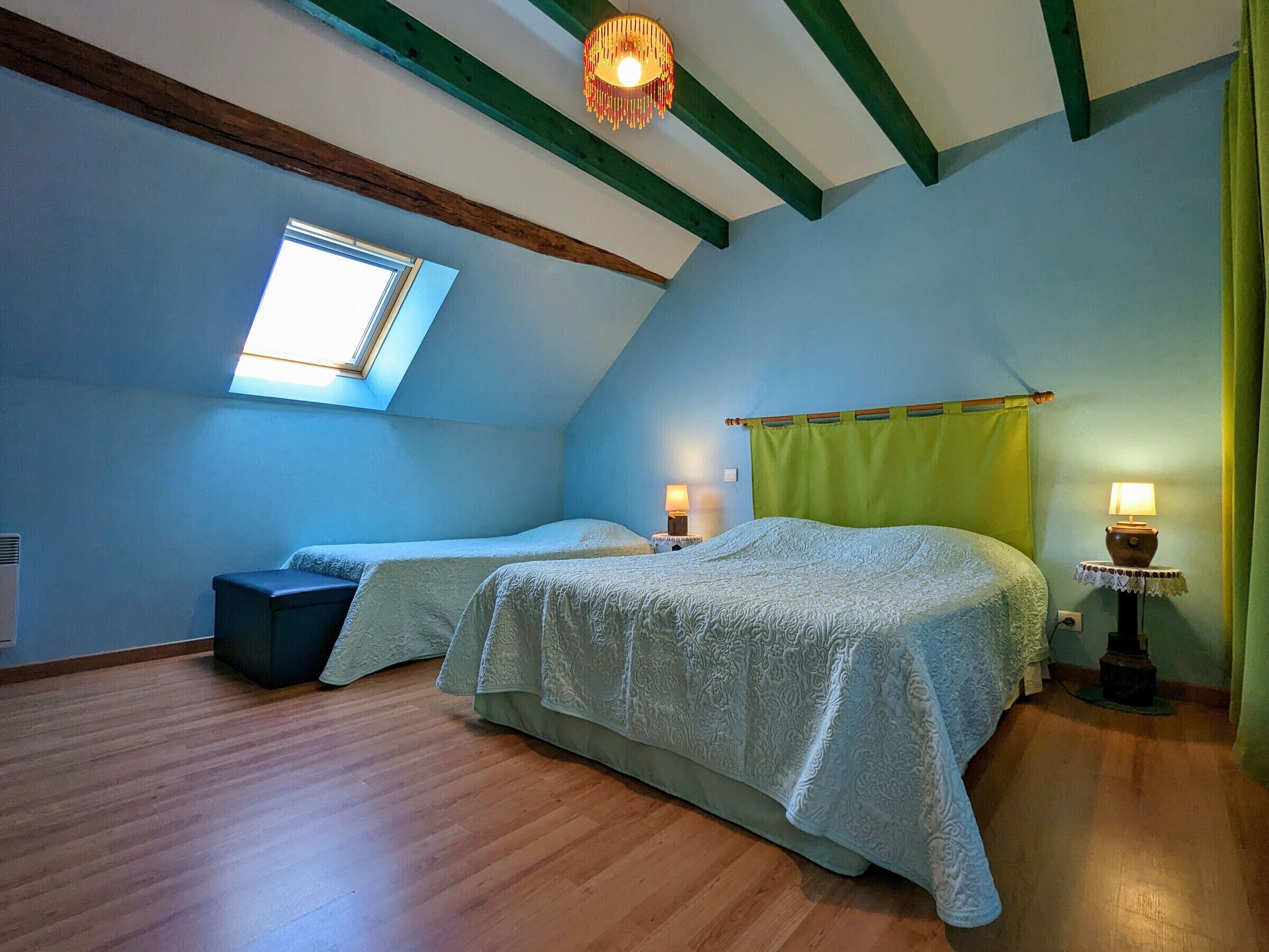A bedroom with two beds, a skylight, wooden beams, and a small table with a lamp.