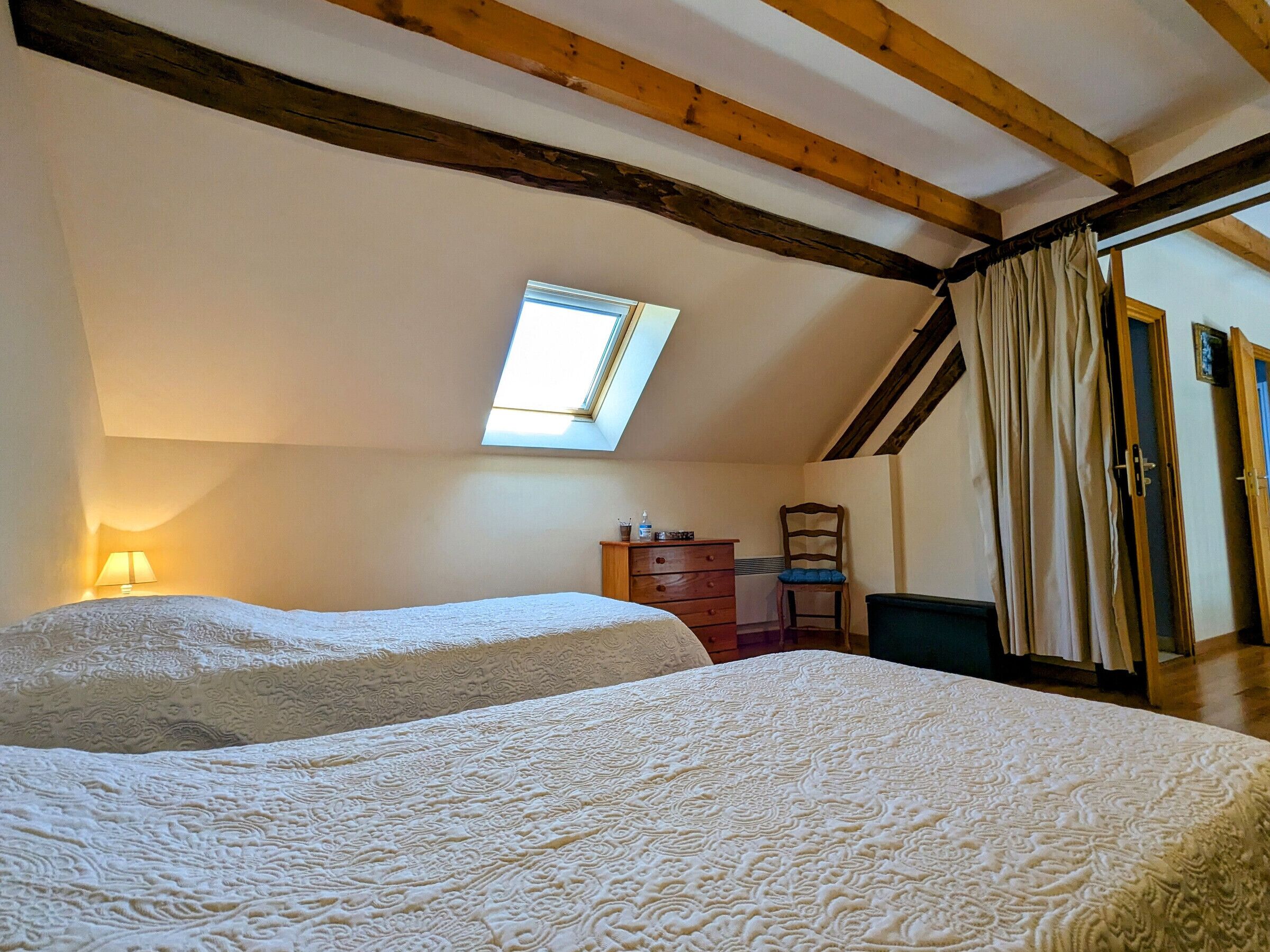 Une chambre avec deux lits, une lucarne, un plafond en bois, une chaise et une commode.