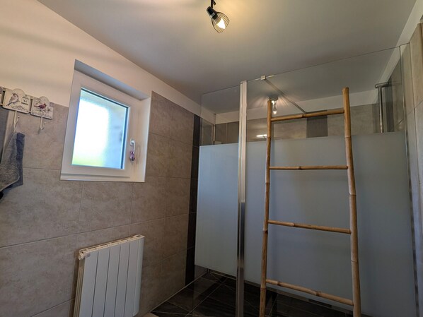 A bathroom with a window, a radiator, a shower with a glass door, and a wooden ladder.