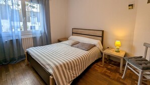A bedroom with a bed, a nightstand, a chair, and a window with curtains. - Charming Country Cottage Near Valençay - 2 Bedrooms, Quiet Garden, Close to Loire Castles & Beauval Zoo (Poulaines)