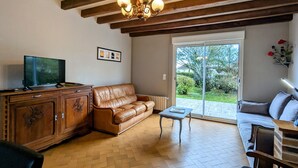 A living room with a wooden ceiling, a leather sofa, a television, a cabinet, and a view through a glass door to the outdoors. - Charming Country Cottage Near Valençay - 2 Bedrooms, Quiet Garden, Close to Loire Castles & Beauval Zoo (Poulaines)