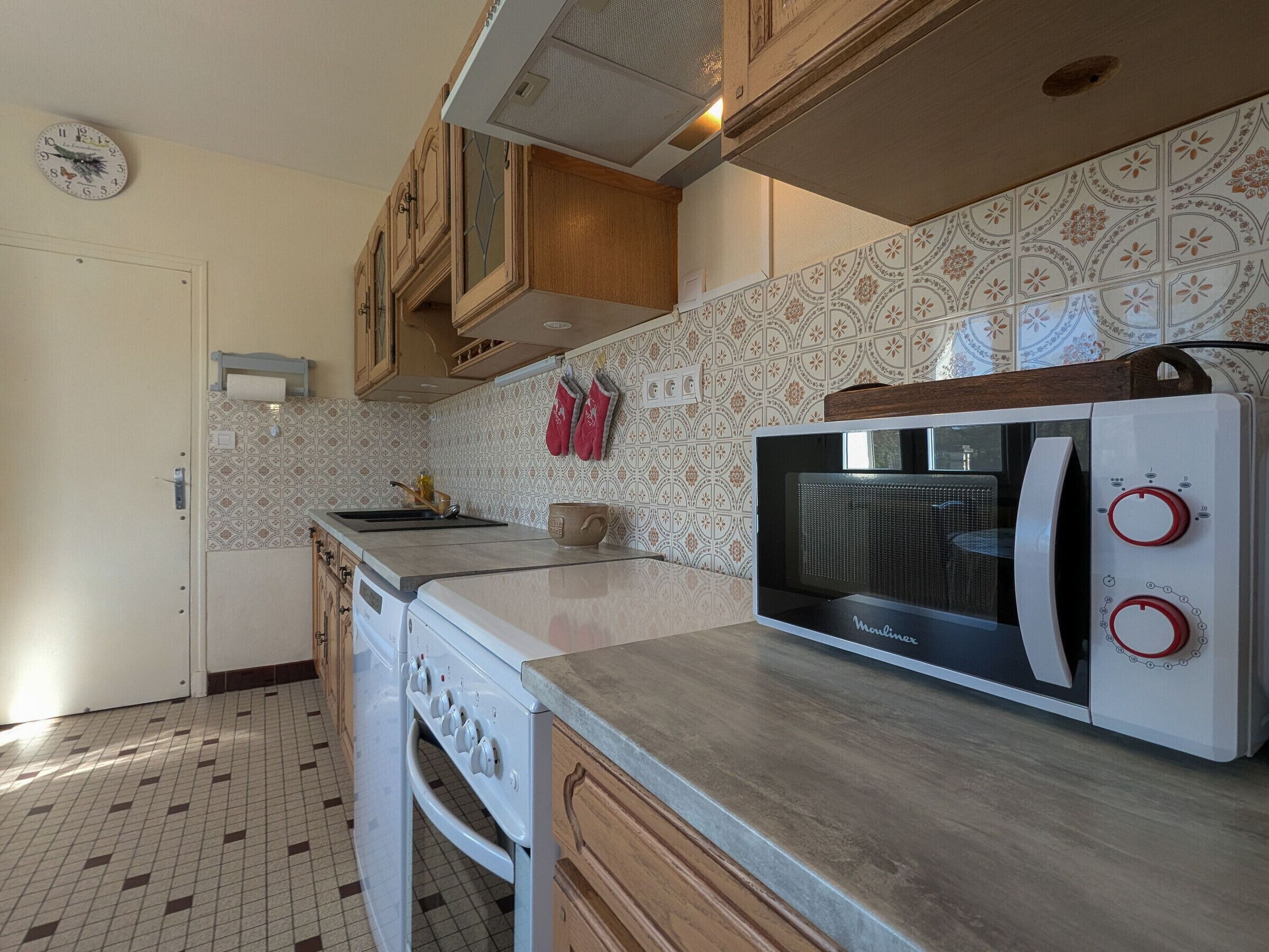 Een keuken met een ingebouwde oven, magnetron en kookplaat. Er zijn houten kastjes en een backsplash met een patroon.