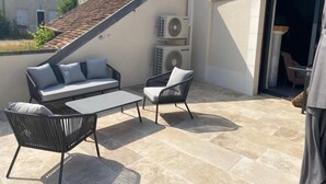 Un patio exterior con un sofá, sillas y una mesa. Se observan equipos de aire acondicionado y una puerta a un área cubierta.