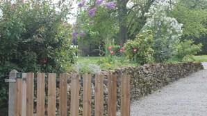 Une clôture en bois, adossée à un mur de pierres, entourée d’un jardin où poussent différentes plantes et fleurs.