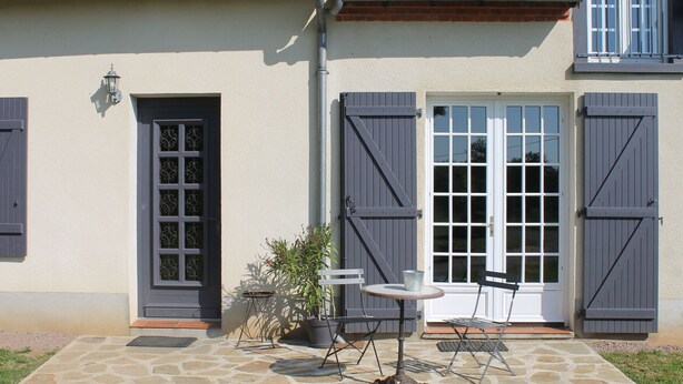 Une maison avec une terrasse en pierre, deux portes avec volets et une petite table et une chaise placées à l’extérieur.