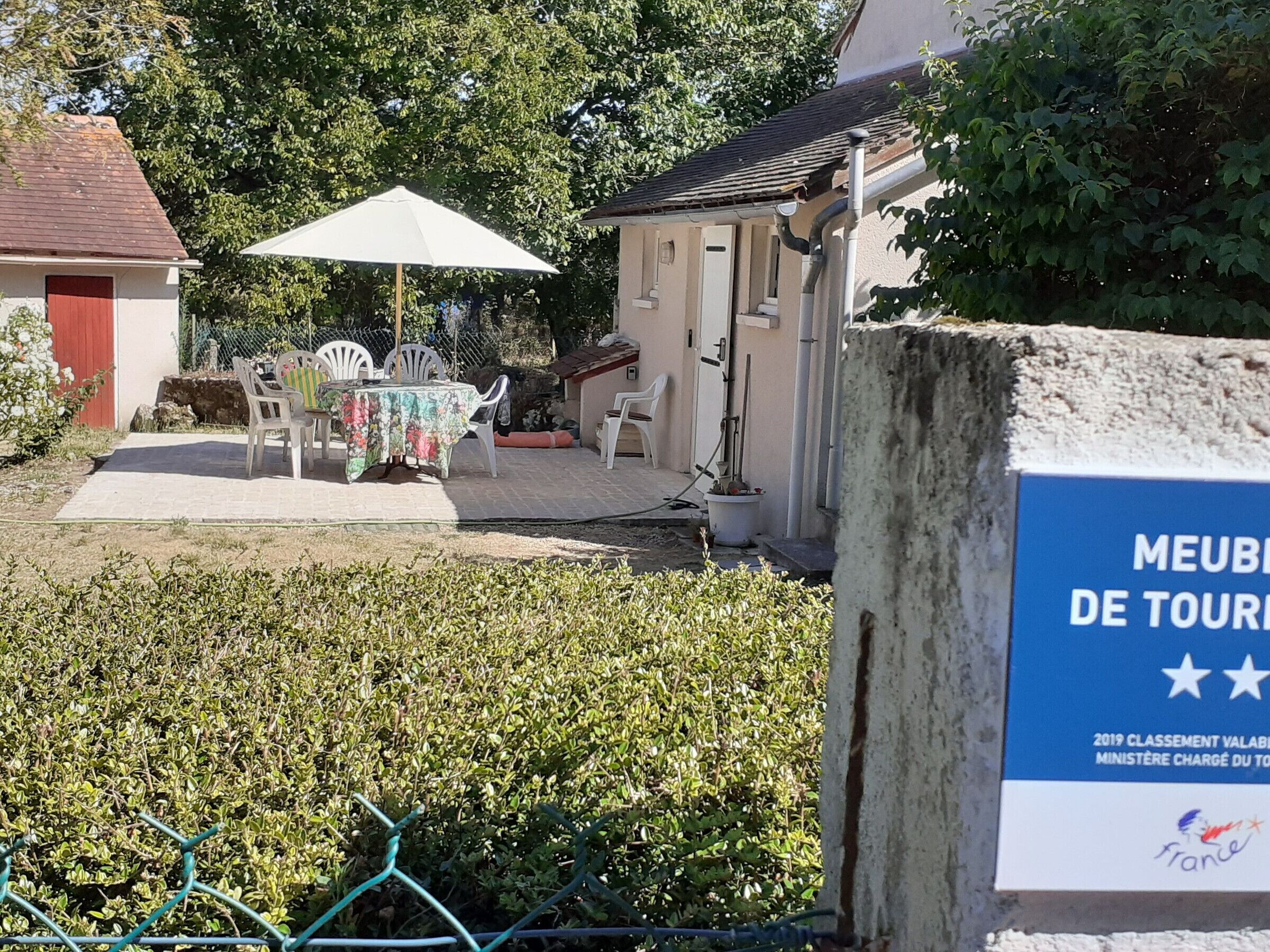 A courtyard with a table and chairs under a white umbrella, surrounded by greenery and a building with a sign reading "MEUBLES DE TOUR".