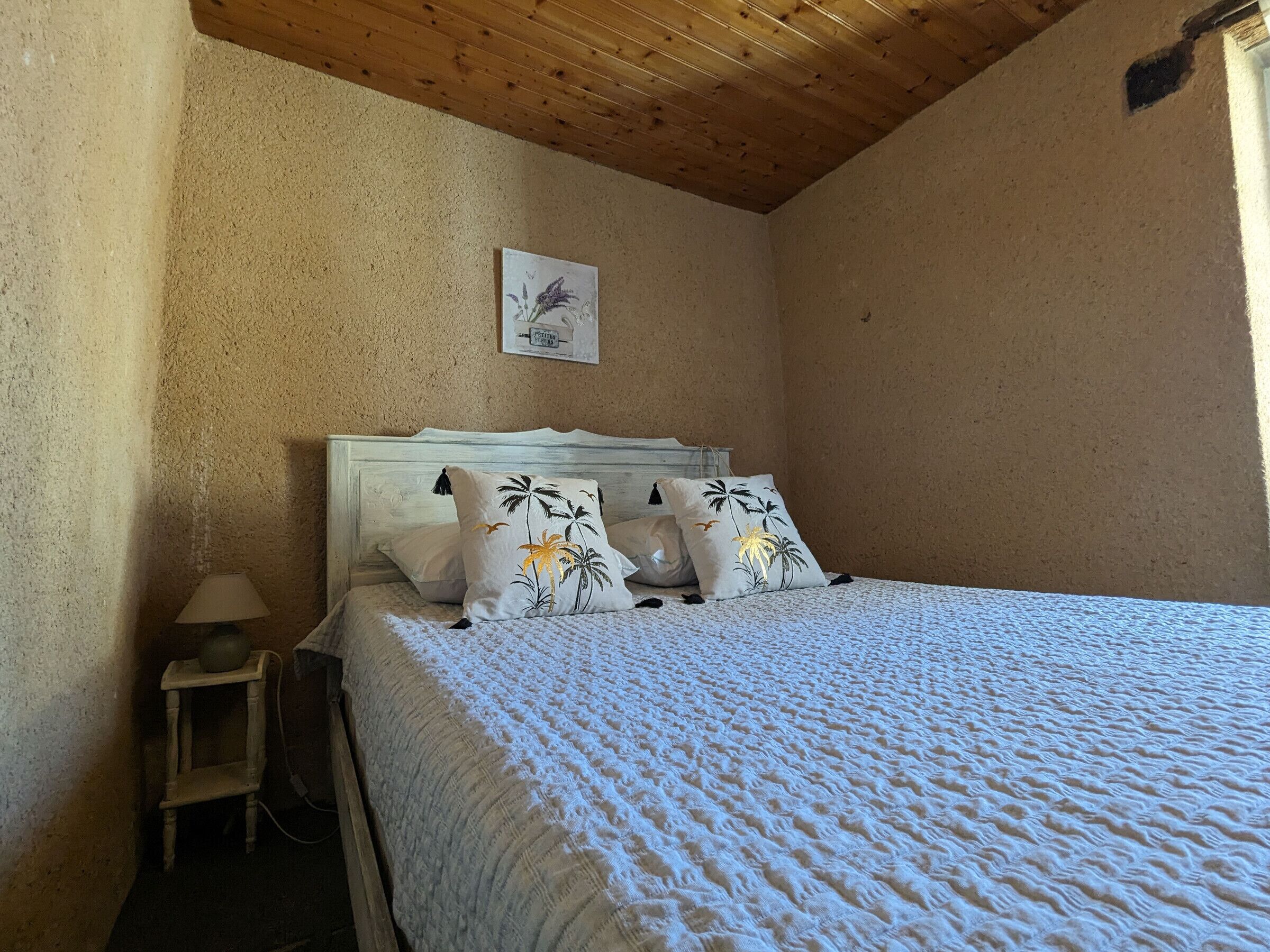 A bedroom with a wooden ceiling, a bed with a white quilt, and a nightstand with a lamp.