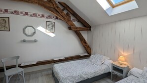 A bedroom with a sloped ceiling, wooden beams, a skylight, a bed with floral bedding, a bedside table with a lamp, and a chair.