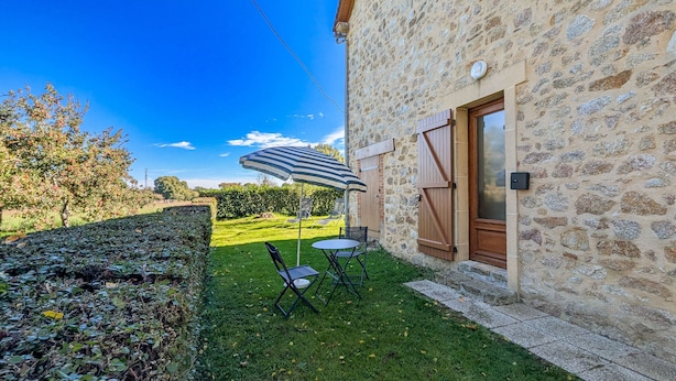 Un bâtiment en pierre avec une porte et des fenêtres, une petite pelouse avec une table et des chaises, et un ciel bleu dégagé.