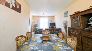 Ein Essbereich mit einer blumigen Tischdecke, Holzstühlen, einem Fernseher und einem Schrank.
