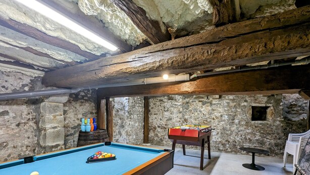 A pool table with balls, a foosball table, and a billiards table in a room with stone walls and wooden beams.