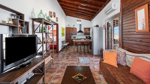 A living room with a TV, wooden furniture, a kitchen area, and a dining table.