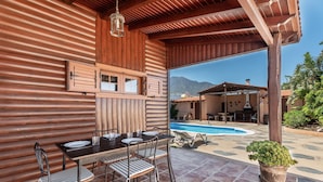A wooden outdoor dining area with a pool and mountains in the background.