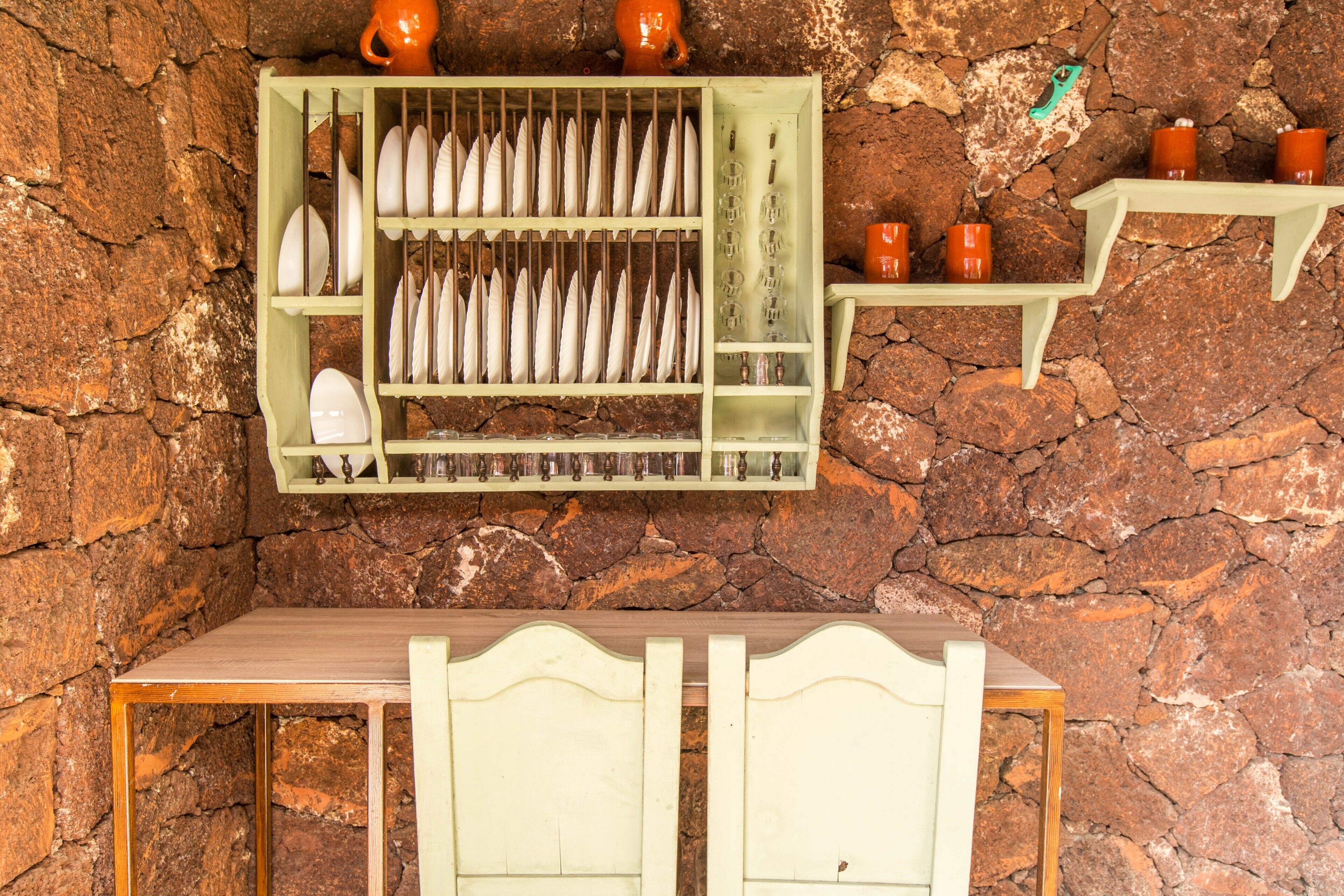 Una cocina rústica con pared de piedra, una mesa de madera y una estantería con platos y tazas.