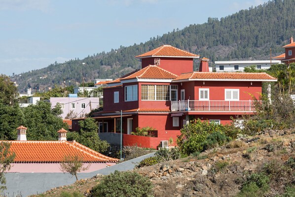 Una casa roja de dos pisos con un balcón, rodeada de vegetación y colinas.