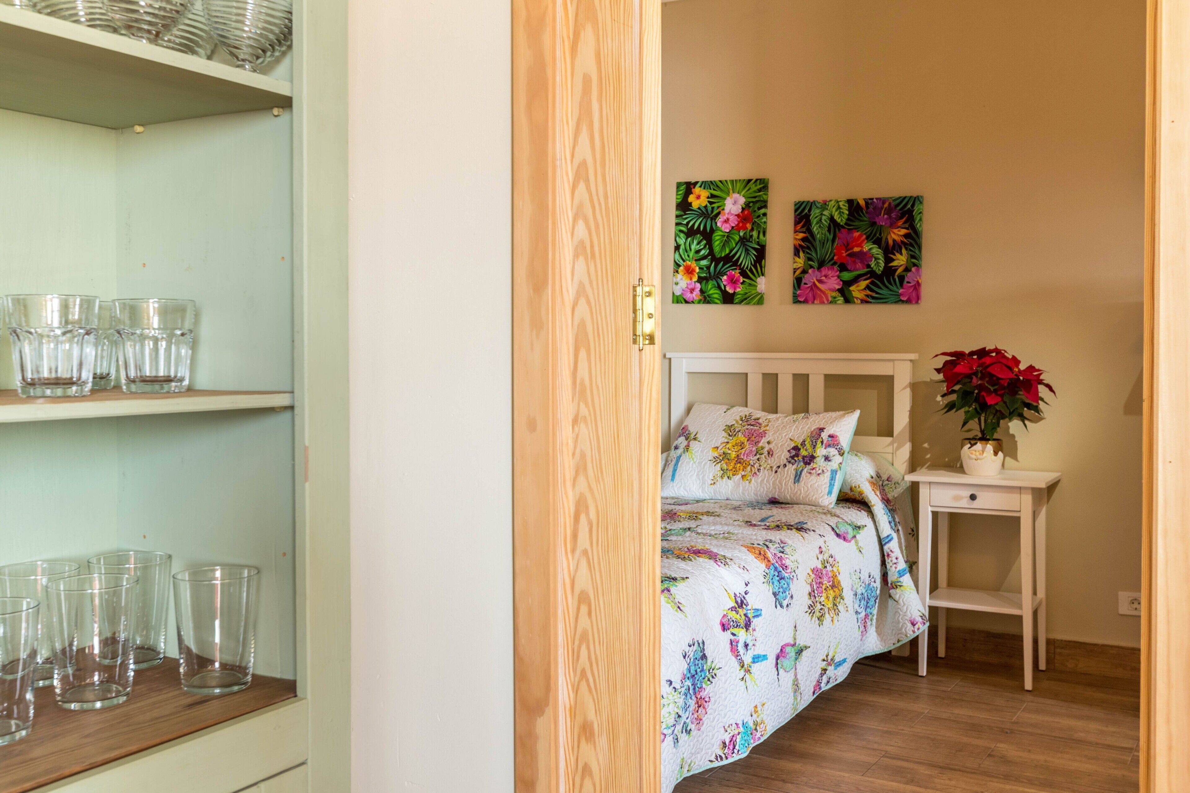 Un dormitorio con una cama, una mesita de noche, una estantería con copas y dos cuadros florales en la pared.