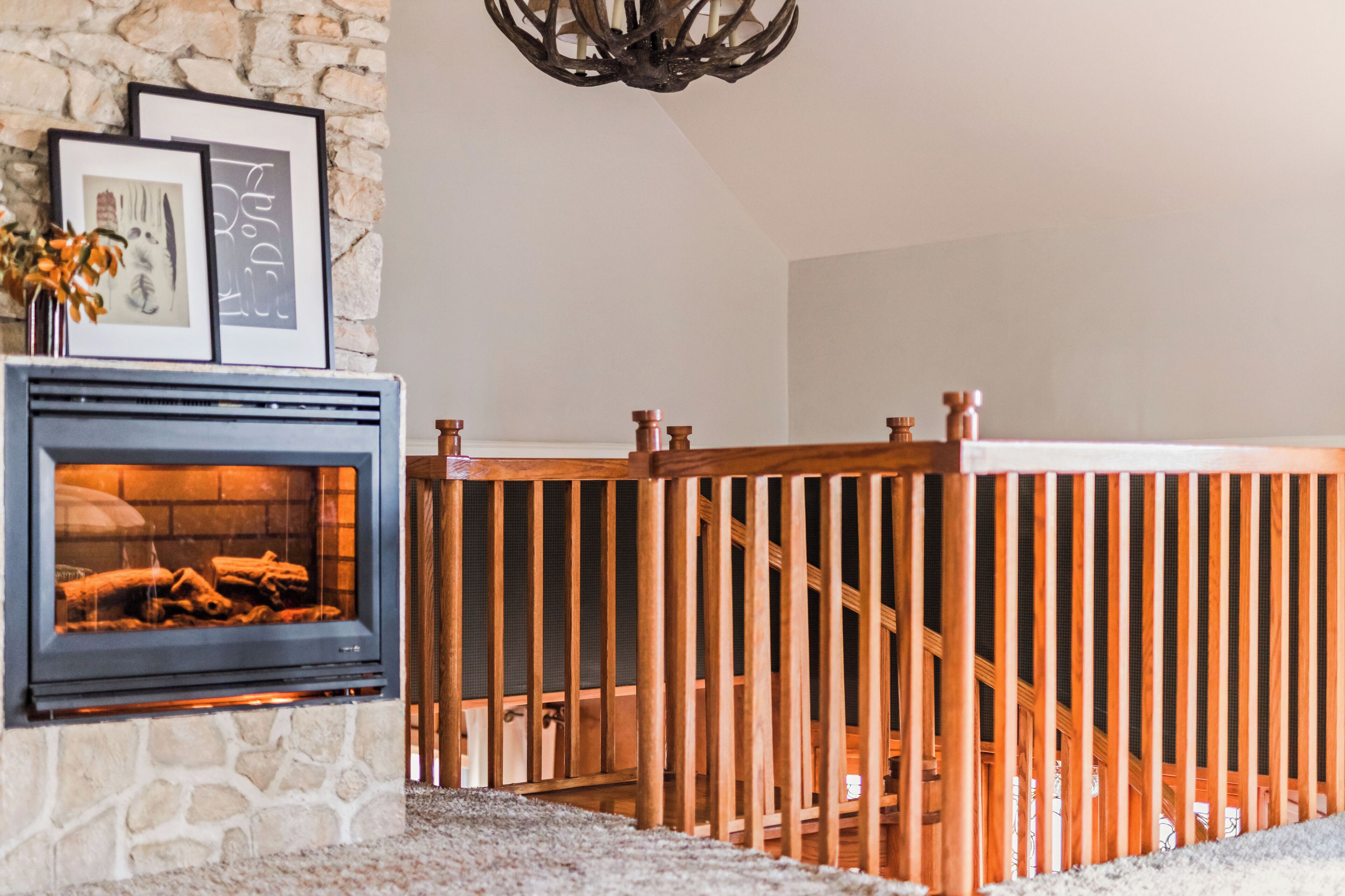 A stone fireplace with framed artwork above it, a wooden staircase railing, and a chandelier.