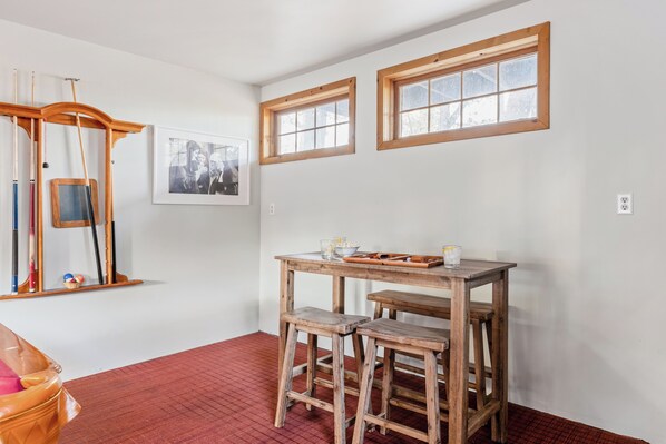 Une pièce avec une table en bois, deux tabourets, une photographie encadrée accrochée au mur et deux fenêtres à cadre en bois.
