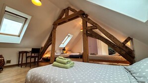 Een zolderkamer met schuine wanden, een dakraam, een bed met een stippenbedekking, een stoel en een bureau met een lamp.