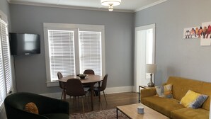 Ein Wohnzimmer mit Sofa, Couchtisch, Esstisch mit Stühlen, einem an der Wand montierten Fernseher und einem Fenster mit Jalousien.