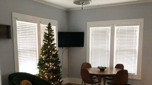 Ein Wohnzimmer mit einem grünen Sessel, einem gelben Sofa, einem Couchtisch, einem Esstisch mit Stühlen, einem an der Wand montierten Fernseher und einem Weihnachtsbaum.