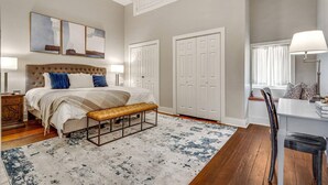 A bedroom with a large bed, a bench, a desk, and a chair. There are two doors and a window with curtains. - Stylish 1BR LGD condo with private balcony on Magazine St. King bed, chef’s kitchen, Wi-Fi, walk to cafés. (New Orleans)