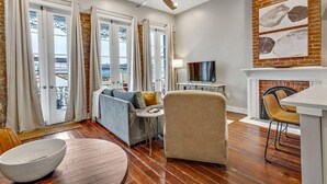 A living room with a sofa, armchair, TV, fireplace, and a balcony with curtains. - Stylish 1BR LGD condo with private balcony on Magazine St. King bed, chef’s kitchen, Wi-Fi, walk to cafés. (New Orleans)