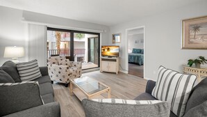 En moderne stue med sofa, lænestol og sofabord i glas. Et fjernsyn er monteret på væggen, og der er en balkon med udsigt.