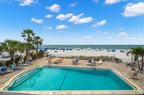 A swimming pool with lounge chairs and palm trees, overlooking a beach with people and umbrellas. - Sandy Shores 203 2 BR Gulf Front (Madeira Beach)