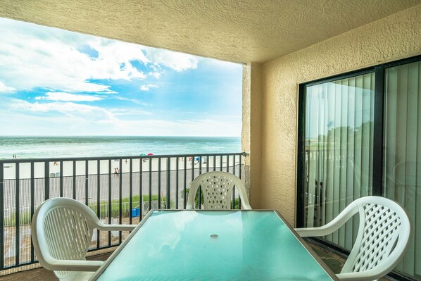 A balcony with a table and chairs, overlooking a beach with people in the distance. - Sandy Shores 203 2 BR Gulf Front (Madeira Beach)