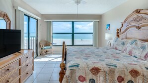 A bedroom with a large bed, a television, a ceiling fan, and a view of the beach. - Sandy Shores 203 2 BR Gulf Front (Madeira Beach)