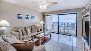 A living room with a sofa, coffee table, and TV, a ceiling fan, and a balcony with a view of the ocean. - Sandy Shores 203 2 BR Gulf Front (Madeira Beach)
