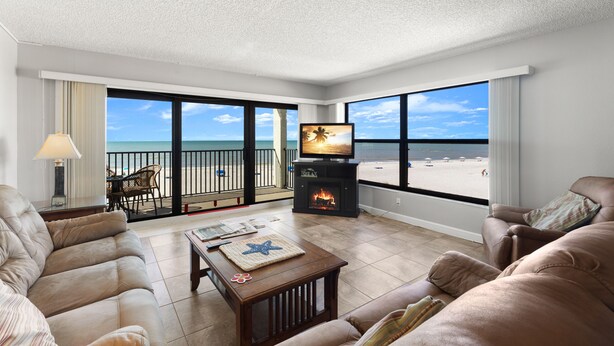 Una sala de estar con chimenea, ventanas amplias con vista a la playa y un balcón con mobiliario exterior.