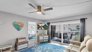 A living room with a sofa, a blue rug, a ceiling fan, a heart-shaped decoration, a television, and a sliding glass door leading to a balcony. - Sandy Shores 311 Water View 2 BR (Madeira Beach)