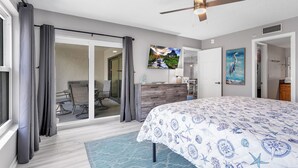 A bedroom with a bed, a ceiling fan, a large window with curtains, a sliding glass door leading to a balcony, and a painting on the wall. - Sandy Shores 311 Water View 2 BR (Madeira Beach)