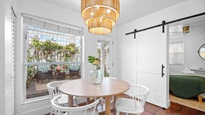 Una habitación con una mesa redonda de madera, sillas blancas, una lámpara colgante, una puerta corrediza y una cama con colcha verde.