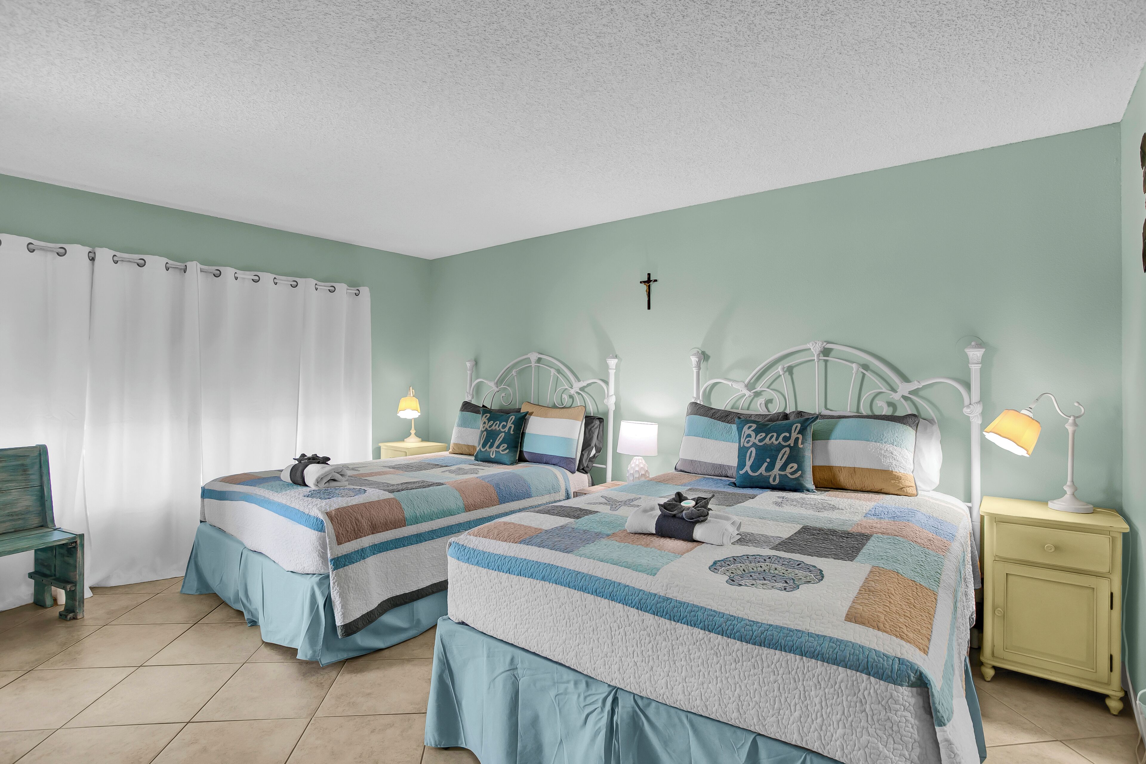A bedroom with two beds, each with a quilted bedspread, a bench, a nightstand with a lamp, and a cross on the wall.