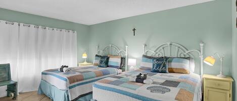 A bedroom with two beds, each with a quilted bedspread, a bench, a nightstand with a lamp, and a cross on the wall.