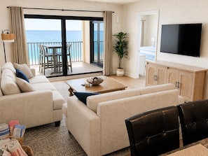 Ein Wohnzimmer mit einer Couch, einem Couchtisch und einem Fernseher. Ein Balkon bietet Blick auf das Meer.