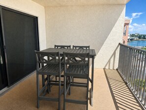 Un balcón con mesa y sillas, vista a un cuerpo de agua y un edificio al fondo.