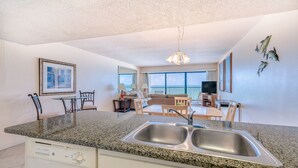 A modern kitchen with granite countertops, a double sink, and a dishwasher. There is a dining area with a table and chairs, and a living room with a sofa and a TV. - Sandy Shores 504 1 BR Gulf Front (Madeira Beach)