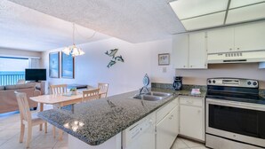 A modern kitchen with granite countertops, a built-in oven, and white cabinetry. There is a dining area with a table and chairs, and a ceiling fan. - Sandy Shores 504 1 BR Gulf Front (Madeira Beach)